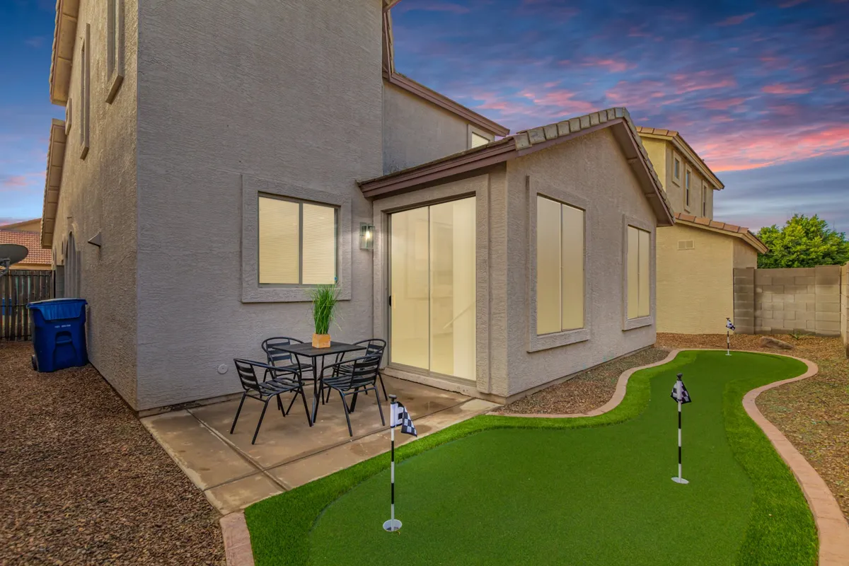 The photo features a backyard putting green adjacent to an outdoor dining set. Warm light spills from home windows, and the scene suggests a space for both golf practice and social gatherings at sunset.