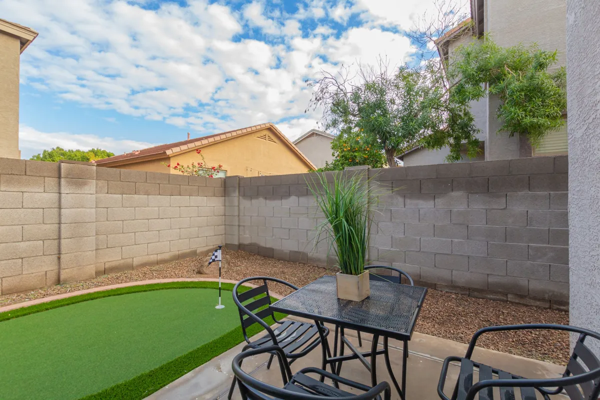 A vibrant backyard scene featuring a miniature golf putting green, a black metal table with chairs for outdoor dining, and a decorative potted plant. The area is bordered by greenery, inviting both play and relaxation.