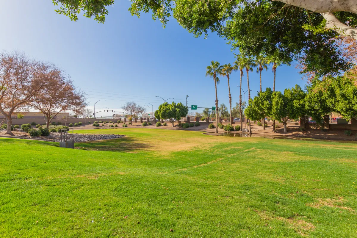 A lush, green park area with palm and deciduous trees, expansive grassy lawns, and a winding walking path—perfect for outdoor games, jogging, or family gatherings in the community.