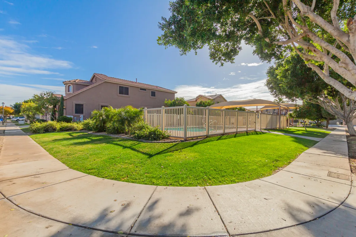 A spacious community pool surrounded by a green lawn and tall trees, all within a fenced area. The inviting pool area is ideal for sunbathing, swimming, or socializing, with space for community events or casual relaxation.