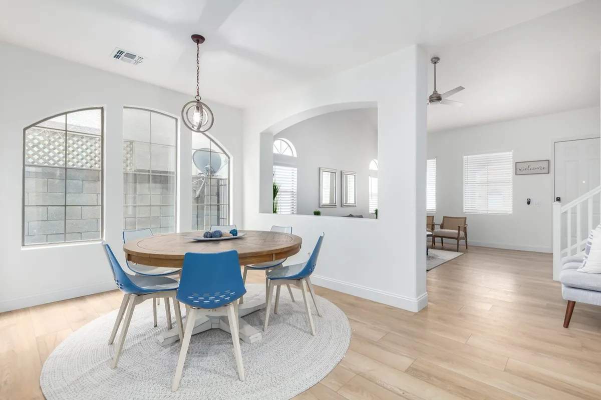 A stylish adjacent dining area featuring a dining table surrounded by blue chairs, large windows that fill the space with natural light, and a decorative light fixture overhead. The ambiance is warm and inviting, ideal for family meals, celebrations, or game nights.