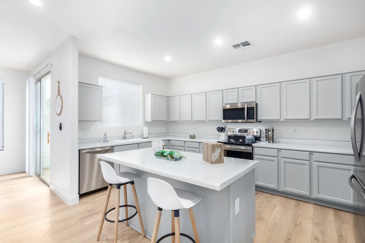 A modern kitchen featuring a central island with seating, brand new stainless steel appliances, and light grey cabinetry. The countertops gleam under ample lighting, and a decorative centerpiece adds a touch of warmth and style. The space is open, inviting, and perfect for both everyday cooking and entertaining guests.