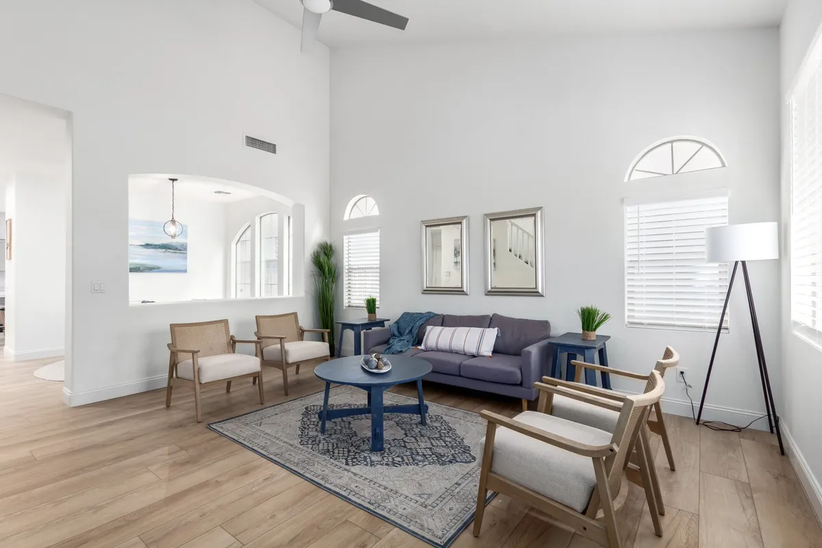 A spacious living area with a sofa, coffee table, and armchairs arranged to encourage conversation and relaxation. Decorative mirrors and a lush plant add a touch of elegance, while large windows flood the room with natural light and offer glimpses of the manicured yard outside. The vaulted ceiling contributes to an airy, open feel.