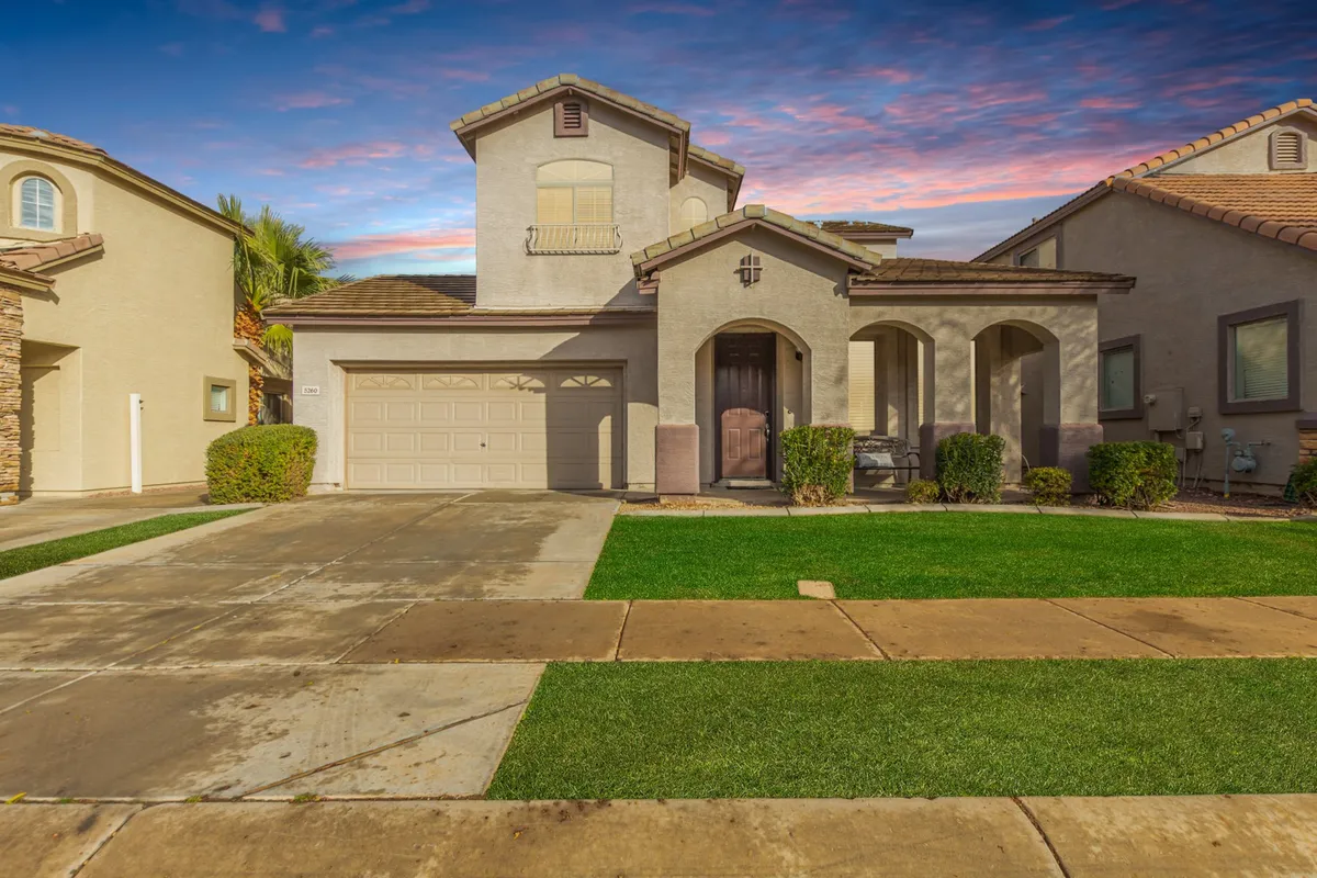 The image showcases the front exterior of a beautifully remodeled single-family house in Mesa, Arizona. The view features a spacious, well-manicured yard with lush landscaping, a welcoming front entrance, and a modern garage door—all bathed in the warm glow of sunset. The inviting porch hints at relaxing evenings and the calm neighborhood surroundings.