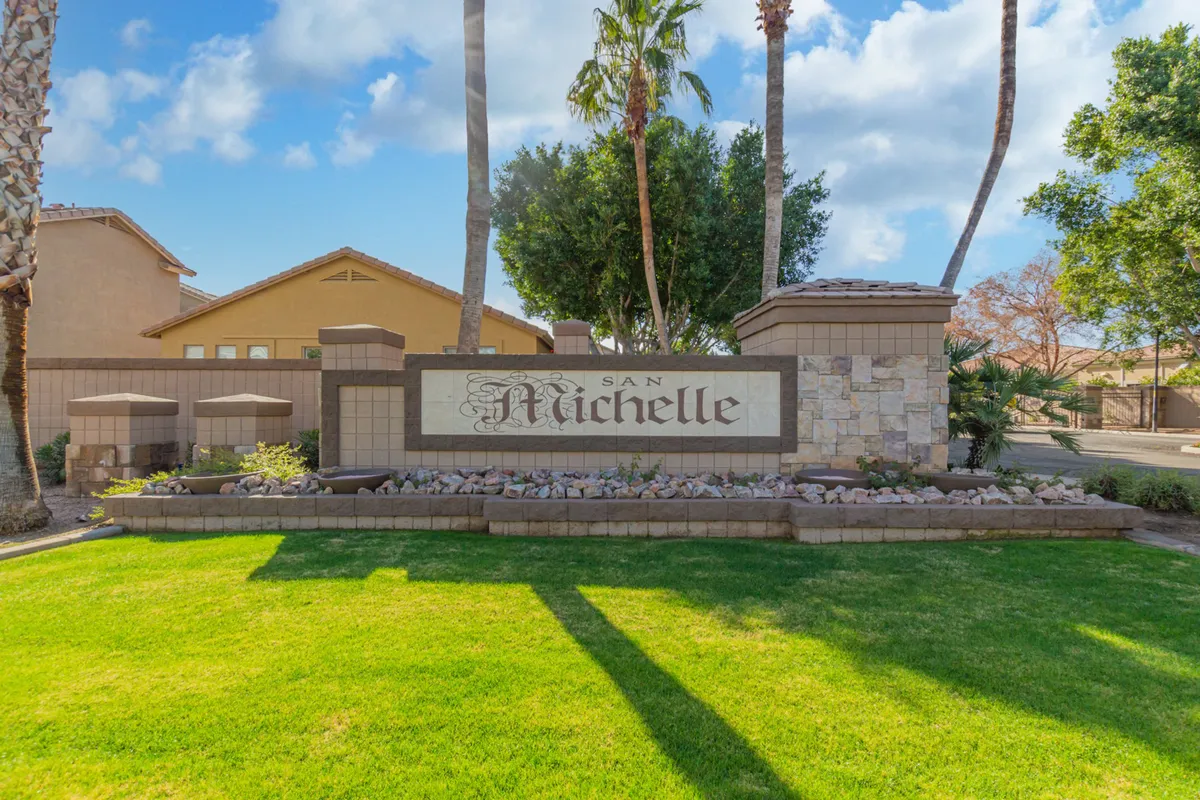 The welcoming entrance to a gated community, highlighted by a stylish property sign, landscaped garden beds, and mature palm trees. The environment exudes serenity and exclusivity, signaling security and community pride.