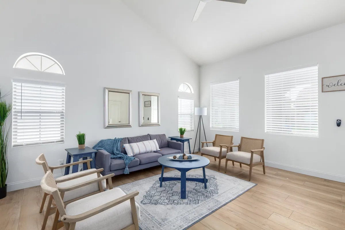 A bright and welcoming living space featuring a comfortable sofa, coffee table, decorative plants, framed mirrors, and expansive windows. The room is awash in natural light, emphasizing the new floors and open-plan design. The layout invites both relaxation and socialization, with sightlines extending to other areas of the home.