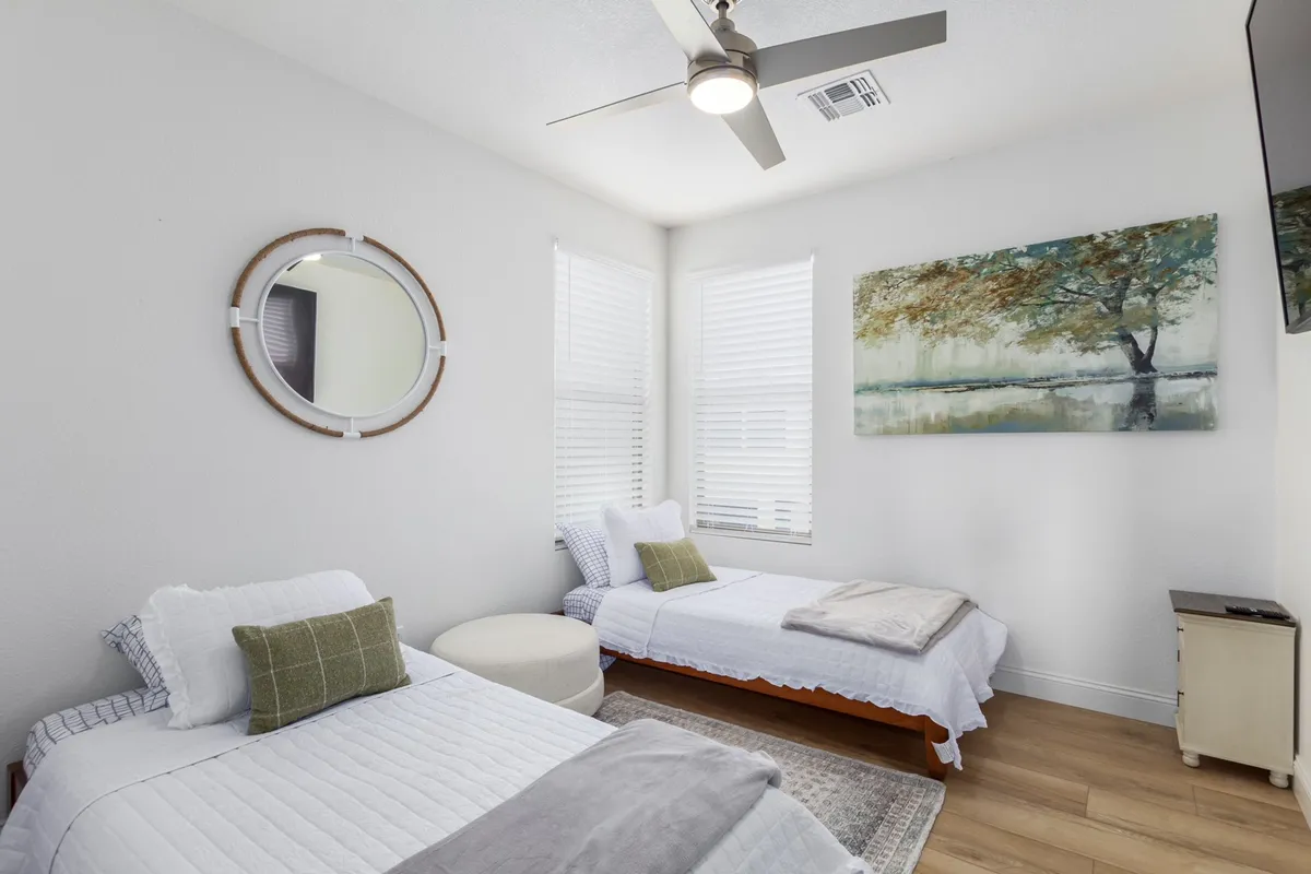 A serene bedroom with two twin beds, a round mirror, a large painting of a tree, a small circular ottoman, and a wall-mounted TV. The room is bathed in natural light, creating a calm and inviting ambiance ideal for relaxation, reading in bed, or unwinding with a favorite show.