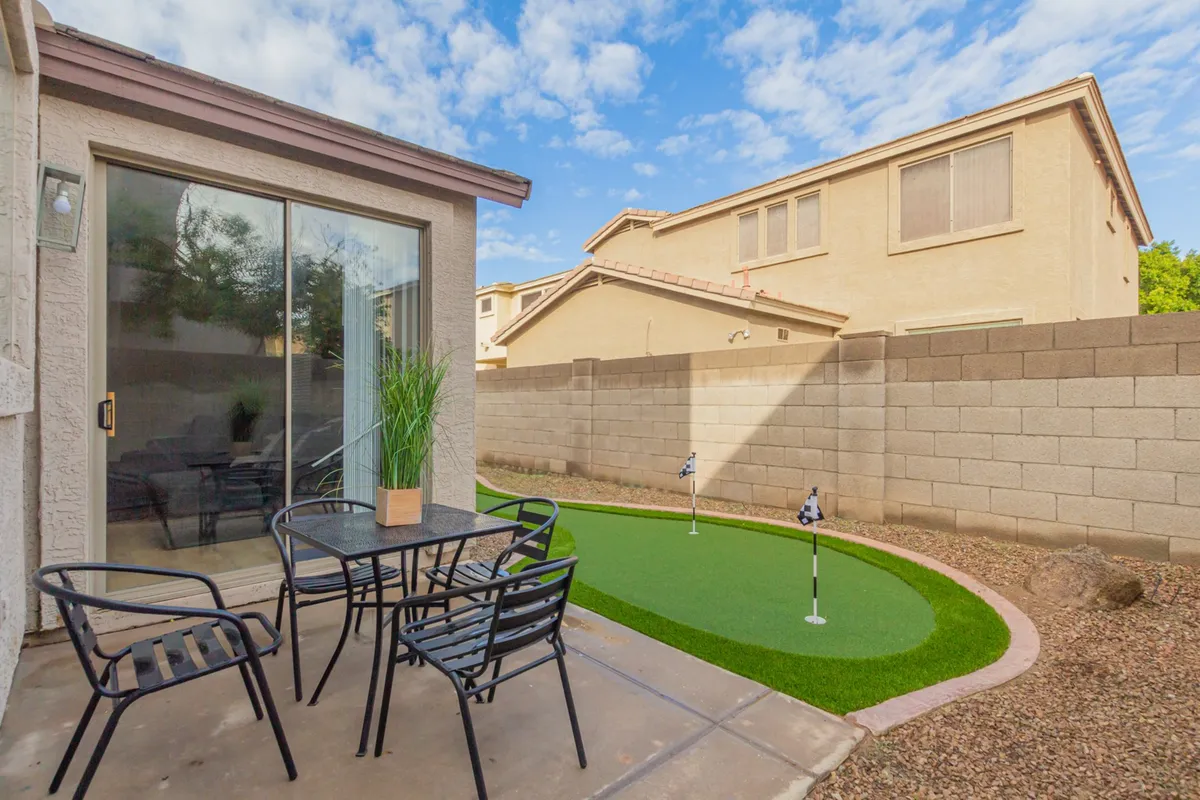 An outdoor space featuring a small putting green and a dining table with chairs, set alongside a glass sliding door that hints at the home's modern interior. The setting is casual yet refined, ideal for relaxation or informal meetings.